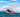 A red and black inflatable boat with one person aboard planes across choppy blue water, its bow lifting high and creating a large wake. This boat makes a perfect flood rescue and emergency response solution.