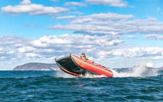 A red and black inflatable boat with one person aboard planes across choppy blue water, its bow lifting high and creating a large wake. This boat makes a perfect flood rescue and emergency response solution.