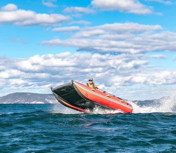 A red and black inflatable boat with one person aboard planes across choppy blue water, its bow lifting high and creating a large wake. This boat makes a perfect flood rescue and emergency response solution.