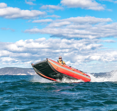 A red and black inflatable boat with one person aboard planes across choppy blue water, its bow lifting high and creating a large wake. This boat makes a perfect flood rescue and emergency response solution.