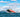 A red and black inflatable boat with one person aboard planes across choppy blue water, its bow lifting high and creating a large wake. This boat makes a perfect flood rescue and emergency response solution.