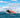 A red and black inflatable boat with one person aboard planes across choppy blue water, its bow lifting high and creating a large wake. This boat makes a perfect flood rescue and emergency response solution.